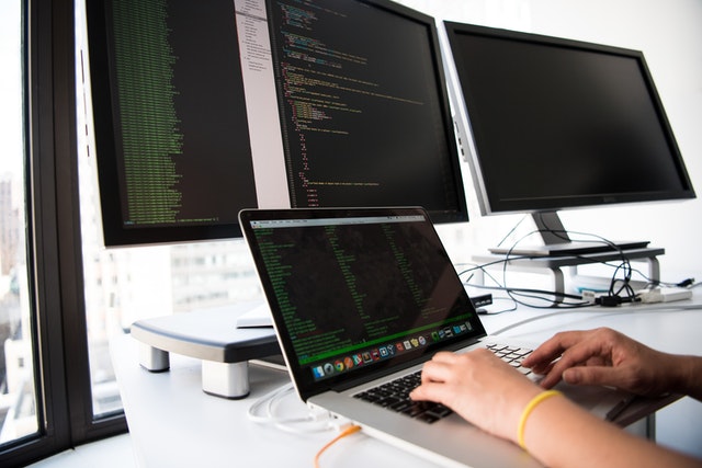 person typing code on a notebook with three screens behind it